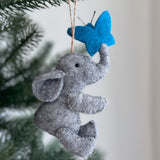 A wool felt elephant with butterfly Christmas ornament hangs on a green Christmas tree branch. The gray elephant features large ears, small black eyes, and a raised trunk. Its trunk gently touches a bright blue butterfly with small black antennae. A brown jute string is attached to the elephant's head for hanging.