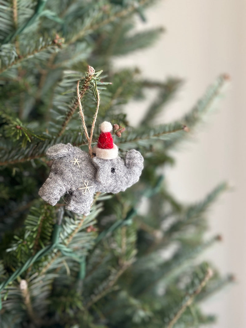 A festive gray wool felt elephant ornament, adorned with delicate snowflake embroidery, hangs from a lush green Christmas tree. A jute string allows for hanging. The blurred white background creates a cozy holiday atmosphere, perfect for Christmas tree ornaments.