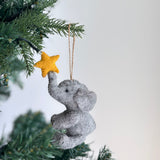 A handcrafted felt elephant ornament with a yellow star hangs from a green Christmas tree branch. The light gray elephant reaches its trunk towards the star, featuring visible textured felt, black eyes, ears, and front legs. It is suspended by a jute string, evoking a joyful and playful mood.
