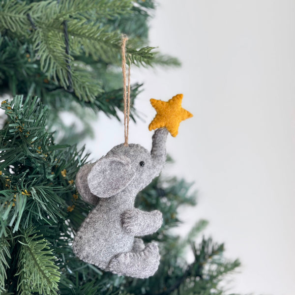 A handcrafted felt elephant ornament featuring a star hangs on a festive Christmas tree. This gray felt elephant is posed in a seated position with its trunk reaching upwards, holding a small yellow star at its tip. A jute string loop is attached for easy hanging, highlighting its unique handmade aesthetic.