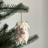 A charming fluffy sheep felt ornament hangs from a green Christmas tree branch, creating a festive atmosphere. This off-white needle-felted wool sheep ornament features a textured body, small pink ears, and a smiling face with a pink nose. It is suspended by a simple jute string.
