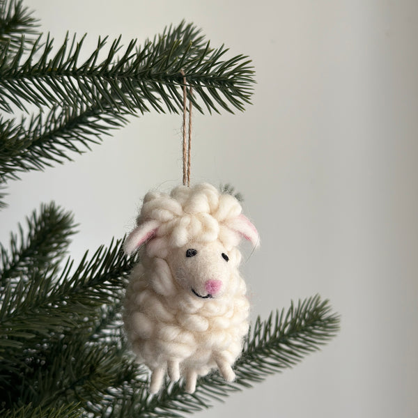 A charming fluffy sheep felt ornament hangs from a green Christmas tree branch, creating a festive atmosphere. This off-white needle-felted wool sheep ornament features a textured body, small pink ears, and a smiling face with a pink nose. It is suspended by a simple jute string.