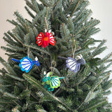 Flying Dragon felt ornaments are playfully displayed on a lush Christmas tree, enhancing the festive decor. These unique, handcrafted 3D needle felted ornaments are made from 100% wool. Variations include red, blue, grey, and green dragons, each detailed with small horns, a tail, and striped wings, hung by natural jute strings.