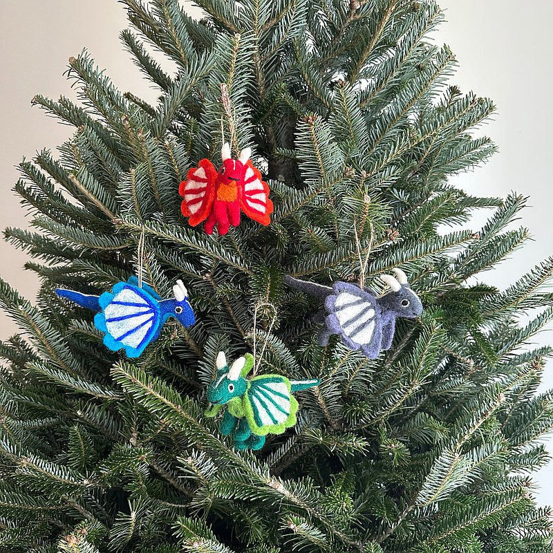 Flying Dragon felt ornaments are playfully displayed on a lush Christmas tree, enhancing the festive decor. These unique, handcrafted 3D needle felted ornaments are made from 100% wool. Variations include red, blue, grey, and green dragons, each detailed with small horns, a tail, and striped wings, hung by natural jute strings.