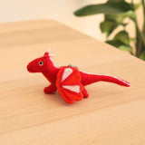 Handcrafted needle-felted flying dragon ornaments are displayed on a light wooden surface with blurred green plants. This whimsical, 3D-designed red dragon has a white stripe from its head to its tail and bold red wings with white stripes. A small metal keychain ring is attached, allowing it to add a touch of fantasy to any space.