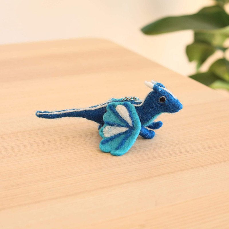 A needle-felted blue flying dragon ornament rests on a light wooden surface, with a blurred green plant in the background, creating a calm, natural atmosphere. This handcrafted 3D ornament is made from 100% wool felt. It features a deep blue body with lighter blue accents on its wings and small white horns, giving it a playful, whimsical appeal.