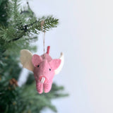 A felt flying pig ornament hangs on a green Christmas tree branch against a bright white background. This charming pink felt ornament features small black eyes, a curly tail, and beige felt wings. It is suspended by a rustic jute string, adding a whimsical touch to the display.