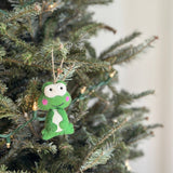 A handcrafted felt frog ornament hangs from a lush green Christmas tree branch, adorned with subtle fairy lights. This charming felt frog features a vibrant green body, bright white eyes, and rosy pink cheeks. Its visibly soft texture adds a whimsical, handmade touch to the holiday decor.