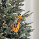 A handcrafted Felt Giraffe Ornament hangs from a green Christmas tree branch adorned with lights. This unique Christmas ornament is primarily yellow-orange with dark brown spots and small black felt ossicones. A jute string is attached, allowing the felt giraffe ornament to hang gracefully, adding safari charm and festive cheer to holiday decor.