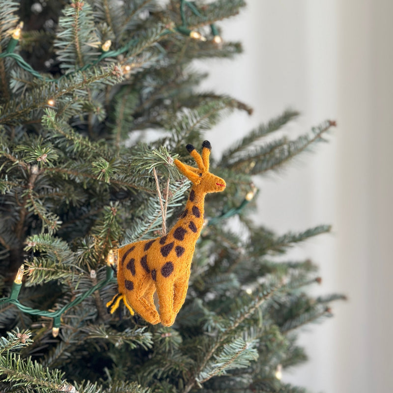 A handcrafted Felt Giraffe Ornament hangs from a green Christmas tree branch adorned with lights. This unique Christmas ornament is primarily yellow-orange with dark brown spots and small black felt ossicones. A jute string is attached, allowing the felt giraffe ornament to hang gracefully, adding safari charm and festive cheer to holiday decor.
