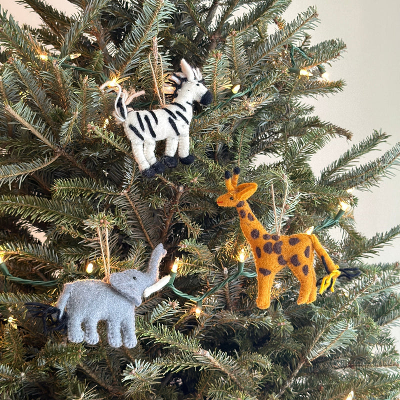 A handcrafted felt giraffe ornament hangs playfully from a Christmas tree branch by a jute string. The unique felt giraffe ornament is textured in orange with distinctive brown spots and features a small yellow tail.