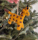 A charming felt giraffe ornament is presented by a hand in front of a Christmas tree. This handcrafted felt giraffe ornament features a yellow body with brown spots, ears, and a tail, showcased against green pine needles and soft white lights. A jute string is attached for hanging.