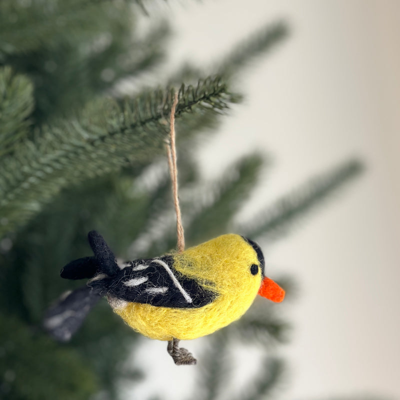 A handmade felt goldfinch ornament hangs on a soft-focused green Christmas tree, adding a playful charm. This delightful Christmas ornament features a plump, bright yellow body with contrasting black and white accents on its wings. Crafted from soft, durable felt, it includes a jute string for easy hanging.
