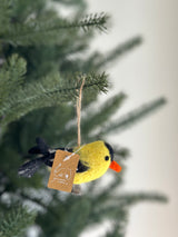 A charming Felt Goldfinch ornament hangs among the green boughs of a Christmas tree, suspended by a jute string. This handmade felt ornament features a bright yellow body and a distinctive black-tipped tail, adding a playful touch to holiday decor.