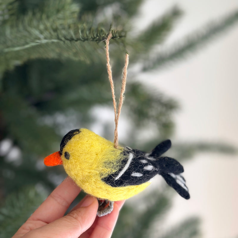 A handmade felt goldfinch ornament is gently held by a female hand, suspended by a jute string against a blurred green pine tree. This charming felt ornament features a bright yellow body, a black cap, and black wings accented with white spots. It has a pointed orange beak and gray felt feet, adding a playful touch.