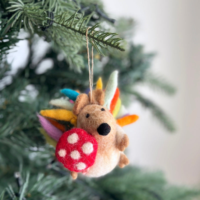 A rainbow wool felt hedgehog Christmas tree ornament hangs on a green branch. This charming hedgehog ornament is crafted from soft brown wool felt with vibrant rainbow-colored spikes. It holds a tiny red mushroom with white spots, and a jute string is attached for easy display.