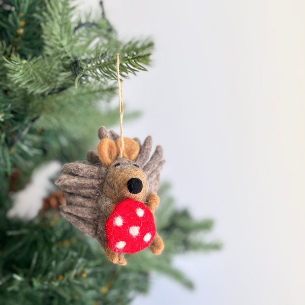 A charming handmade wool felt hedgehog ornament hangs on a Christmas tree. The light brown hedgehog has a textured body with protruding spines, a tan face, and small ears. It holds a red mushroom with white dots, suspended by a jute string. The scene creates a festive, cozy atmosphere against blurred green pine needles.