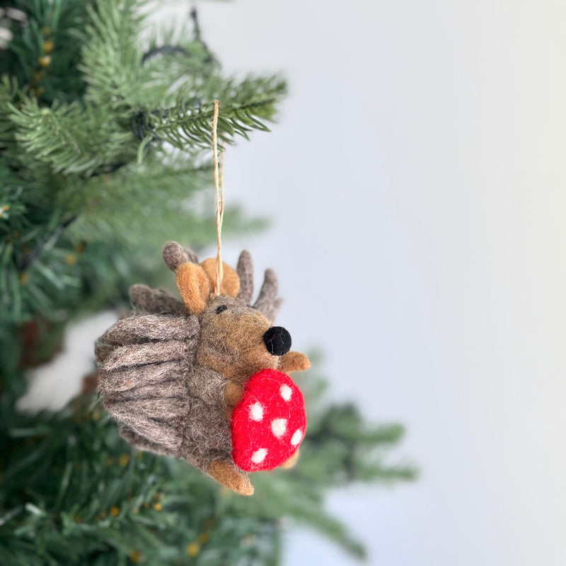A charming wool felt hedgehog ornament hangs from a Christmas tree branch, suspended by a jute string against a soft white backdrop. The handcrafted hedgehog features a textured body, brown ears and limbs, and a black nose. It playfully holds a red mushroom with white spots, adding a whimsical touch to the Christmas tree.