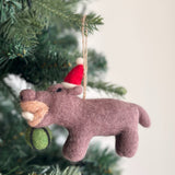 A mauve felt hippo ornament, shown eating a green watermelon, hangs on a festive Christmas tree branch. This handcrafted felt hippo ornament features a lighter tan muzzle with white felt teeth and a red Santa hat with a white pompom. A jute string on its head allows this unique Christmas tree ornament to hang gracefully.
