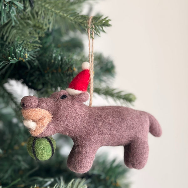 A mauve felt hippo ornament, shown eating a green watermelon, hangs on a festive Christmas tree branch. This handcrafted felt hippo ornament features a lighter tan muzzle with white felt teeth and a red Santa hat with a white pompom. A jute string on its head allows this unique Christmas tree ornament to hang gracefully.