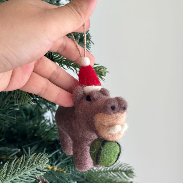 A handcrafted brown wool felt hippo ornament, depicted eating a watermelon, is presented by a female hand against a festive green Christmas tree backdrop. The unique felt hippo ornament features a lighter tan snout, rounded ears, and a red Santa hat with a white brim and pompom. A jute string is attached for hanging this charming Christmas tree ornament.