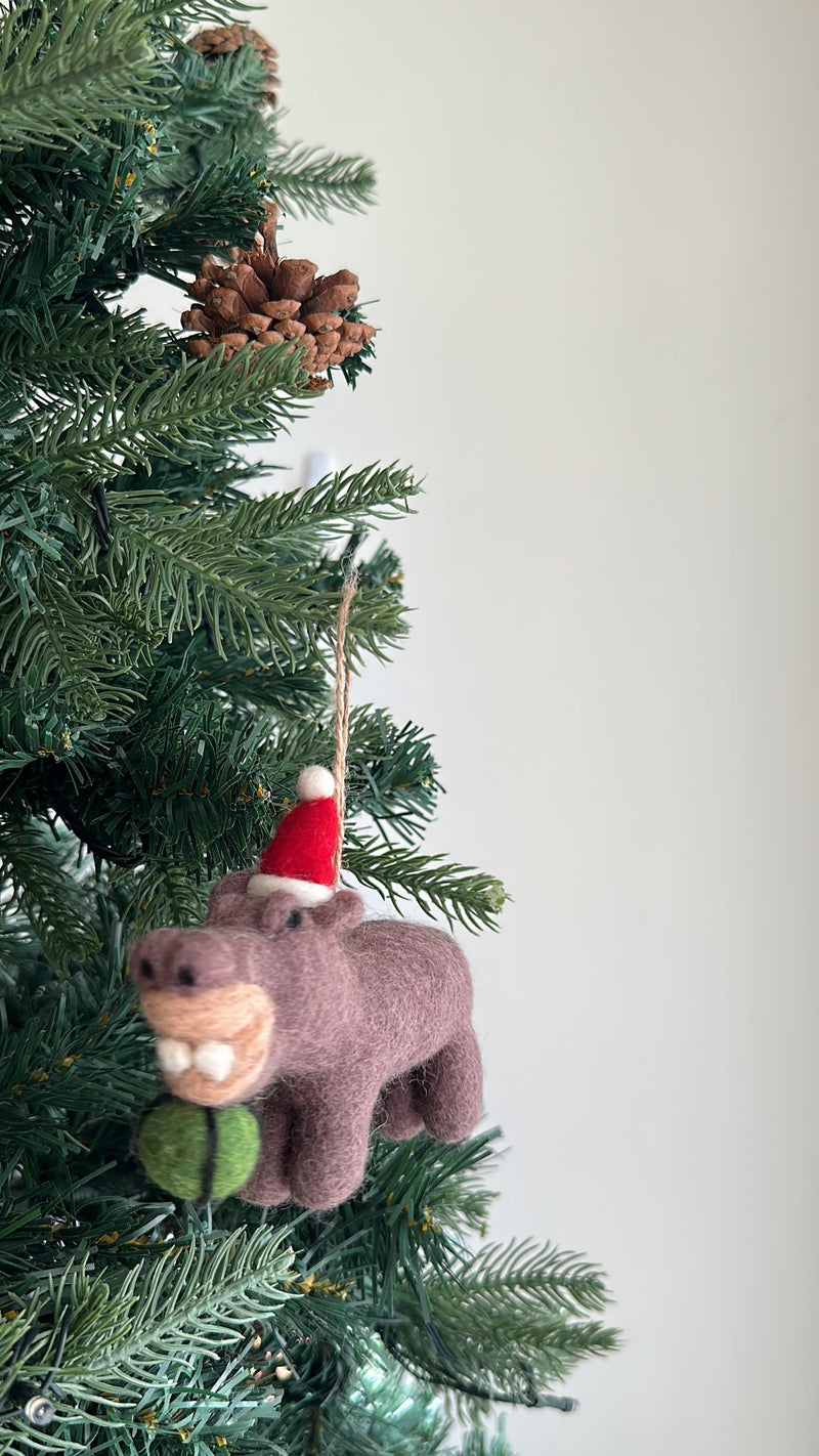 A wool felt hippo ornament, happily eating a watermelon, hangs from a lush green Christmas tree branch. This heathered-brown handcrafted felt ornament has a smiling expression, a tan muzzle, and two white teeth. It wears a festive red hat with a white pompom and holds a green felt watermelon slice with a black stitched line. A brown jute string loop on its back provides a hanger for display.