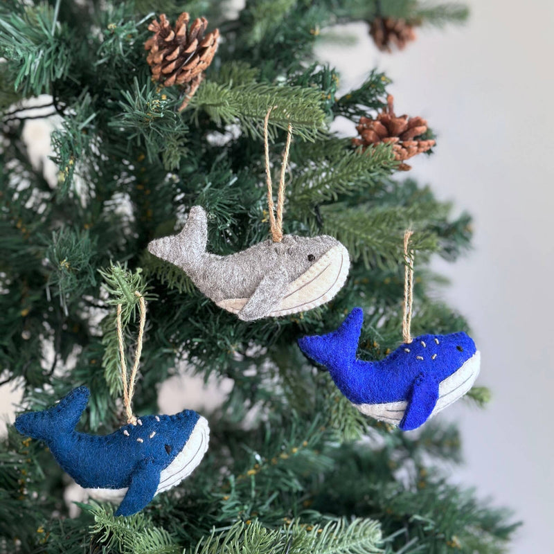 A felt humpback whale ornament hangs on a Christmas tree adorned with pine cones, set against a soft, neutral background. This unique wool felt humpback whale ornament features a light gray body and a white underbelly, with tiny white embroidered details that highlight its needle-felted, hand-stitched texture. It has a small tail and flippers, with a jute string attached for hanging. The handcrafted ornament adds a cozy, festive charm.