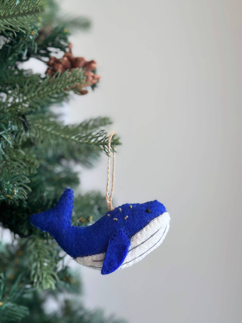 A wool felt Humpback Whale Christmas ornament hangs from a Christmas tree branch, surrounded by green needles and brown pine cones. This charming needle felted ornament, crafted from textured gray wool felt, features a white underside with thin gray lines. It is suspended by a jute string, adding a unique touch to festive decor.