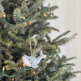 A gray wool felt Humpback whale ornament hangs on a Christmas tree, adorned with small, warm lights against a neutral wall. Hand-stitched from organic wool, this unique Christmas ornament features a lighter underside and a small, round eye. A jute string allows this charming felt ornament to hang gracefully, adding a cozy, festive touch to holiday decor.
