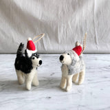 A pair of handcrafted husky felt ornaments with Christmas hats rests on a soft, off-white fabric and a marbled surface. One husky felt ornament features a black and white coat, while the other is gray and white. Each wears a festive red Christmas hat trimmed with white, topped with a jute string for hanging.