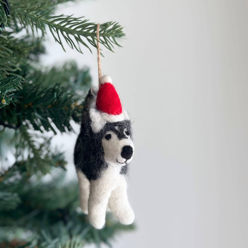 A handcrafted felt husky ornament with a red Christmas hat and white trim hangs by a jute string on a festive Christmas tree. This needle felted husky is crafted from shades of grey and white felt, showcasing its fluffy texture and distinctive markings.