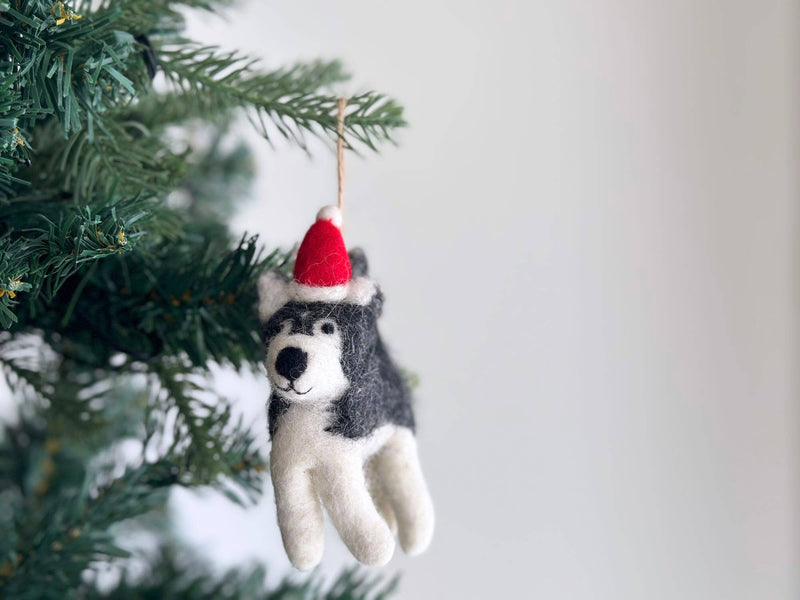 A charming handmade felt husky ornament, adorned with a bright red Christmas hat, hangs on a green Christmas tree branch against a plain white background. The festive husky ornament features a fluffy white belly and paws, contrasted by grey felt on its back and head, with a black nose and simple stitched eyes.