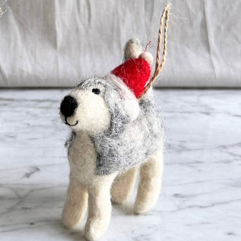 A handcrafted felt husky ornament, ready to hang, is displayed against a light, airy backdrop. This charming Christmas tree ornament features a fuzzy, gray and white husky wearing a bright red Christmas hat adorned with a fluffy white pompom, adding a whimsical touch to holiday decor.