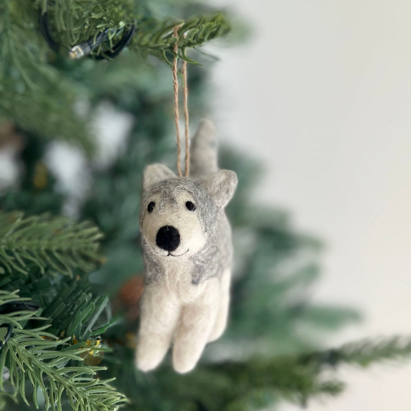 A handcrafted husky felt ornament hangs gracefully from a lush green Christmas tree branch. This unique felt ornament features a blend of gray and white felt, accurately capturing the husky's coat, complete with pointed ears, a black nose, and a bushy tail. It is suspended by a jute string, adding a touch of handmade elegance to the festive decor.