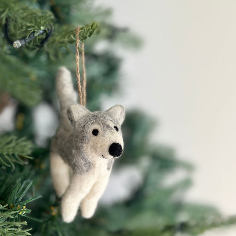 A grey and white husky felt ornament hangs gracefully from a Christmas tree branch, creating a festive atmosphere. This handmade felt husky ornament features perky ears, a tail, and contrasting black eyes and nose, giving it a playful expression. A jute string is attached, ready for hanging.