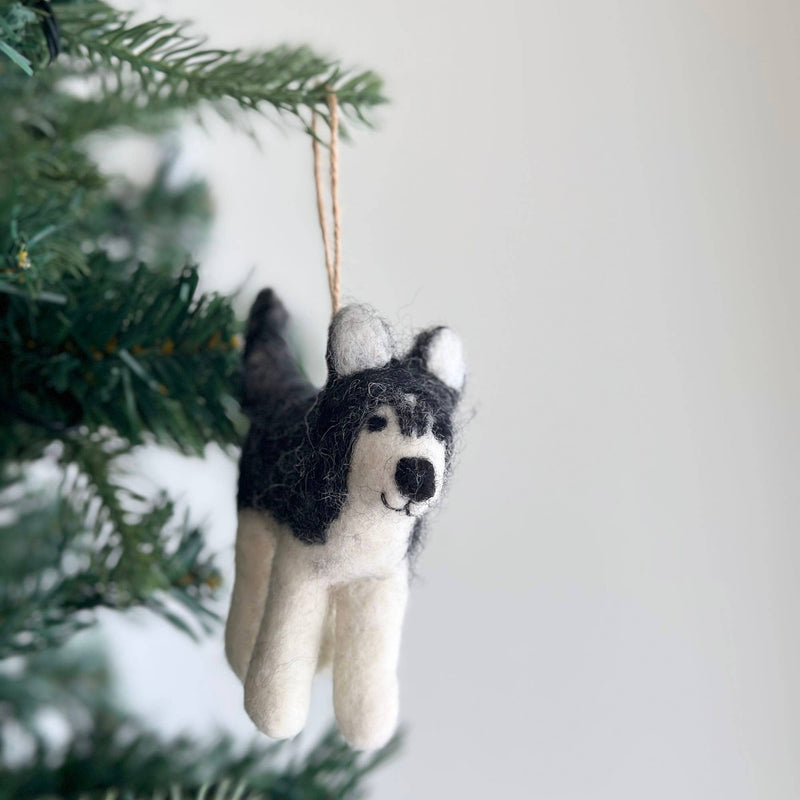 A handcrafted wool felt husky ornament is playfully hung on a Christmas tree branch, set against a simple white background. This black and white husky felt ornament captures distinct markings, featuring upright ears and a smiling face. A jute string connects it to the branch, highlighting its soft, inviting texture and cheerful appearance.