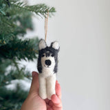 A handcrafted felt husky ornament is displayed near blurred green Christmas tree branches and a plain white wall, evoking a cozy holiday setting. The felt husky ornament is predominantly white with black markings around its head and ears, featuring a soft, textured finish that mimics fur. It has small, perky ears and a sweet expression, with a jute string attached for hanging.