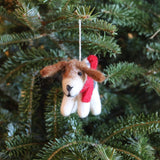A handmade felt Jack Russell dog ornament hangs from a vibrant green Christmas tree branch by a white cotton string. This charming needle felted ornament features a white body, a black nose, and brown ears, with a red scarf tied around its neck.