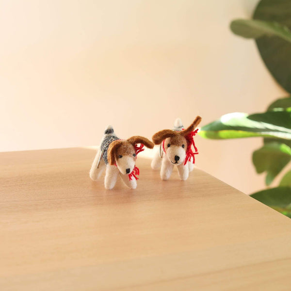 Two handmade Jack Russell dog felt ornaments are displayed on a light wooden surface against a bright, airy backdrop with greenery. These 3D wool felt ornaments, crafted with needle felting, feature white bodies, brown floppy ears, and distinct markings. One ornament has a grey back, while the other sports a decorative red cotton string. These charming figures stand freely, perfect as unique Christmas tree ornaments.