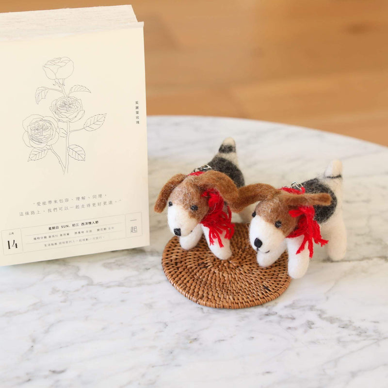 Two wool felt Jack Russell dog ornaments are displayed on a woven mat set on a white marble table, with a book featuring a botanical rose illustration in the background. These handmade ornaments have brown and white bodies with floppy brown ears. Each features a festive red cotton string around its neck and a metal ring on its back, highlighting their artisanal charm.