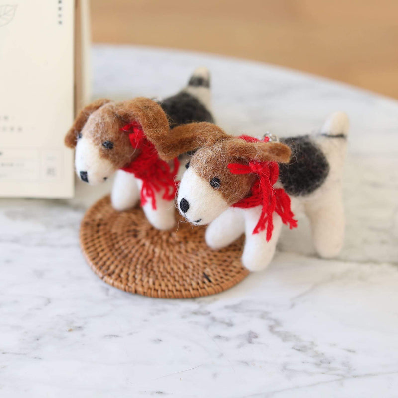 A playful felt Jack Russell dog ornament is showcased on a rustic woven mat, against a bright, airy background. This adorable 3D wool felt Jack Russell dog ornament is expertly crafted in white, brown, and black, featuring small black eyes and a red cotton string around its neck, highlighting its handmade charm.