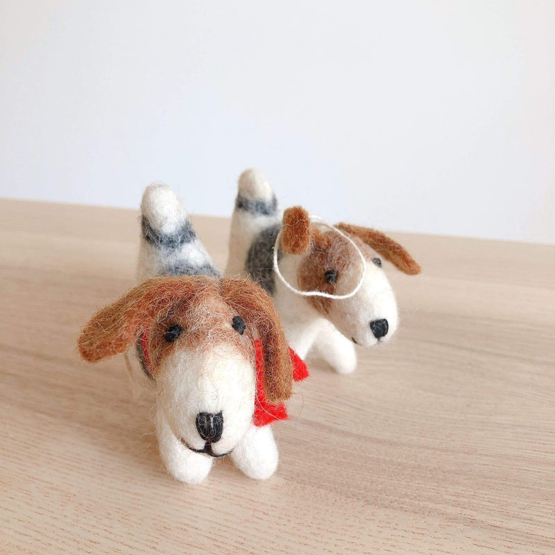 Two handmade 3D felt Jack Russell dog ornaments rest on a light wood surface, evoking a playful and handcrafted feel. Each organic wool Jack Russell dog felt ornament features brown floppy ears, black eyes, and a black nose. One dog wears a red collar, while the other has a white cotton string for hanging, designed for a whimsical Christmas theme.