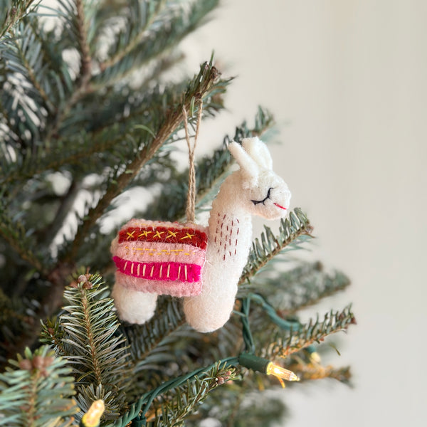 A white wool felt llama ornament hangs on a green Christmas tree, creating a joyful, decorative atmosphere. This handmade llama felt ornament features a long neck, upright ears, a stitched black eye, and a red smile. It is adorned with a colorful saddle blanket in pink, red, and yellow, and has a brown jute string for hanging. The unique Christmas ornament is nestled among pine needles and small, warm-toned lights.