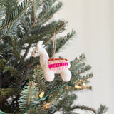 A white wool felt llama ornament hangs gently on a festive Christmas tree, nestled among green pine needles and soft glowing lights. The charming llama features a fluffy texture, stitched details for its eyes, and a colorful striped saddle in brown, yellow, light pink, and hot pink, with a brown jute string for hanging.