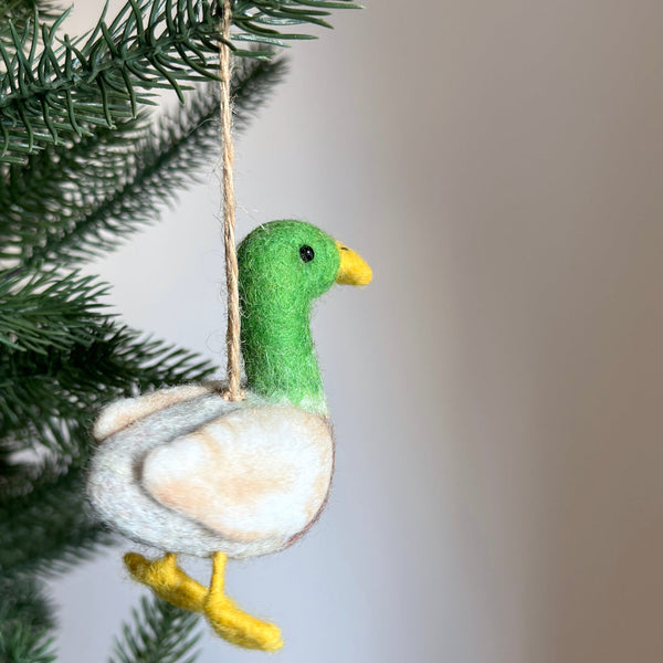 A handcrafted wool felt Mallard Duck ornament hangs from a green Christmas tree branch against a soft grey background. This vibrant felt ornament features a green head, yellow beak, chestnut chest, and signature blue wing patches, with yellow feet dangling below. A jute string is visible for hanging.