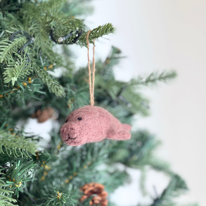 A handcrafted felt manatee ornament is displayed on the green branches of a Christmas tree. This needle felted manatee ornament has a soft, rounded pink body with black eyes and a stitched mouth. A jute string is attached for easy hanging, adding a whimsical touch to holiday decor.