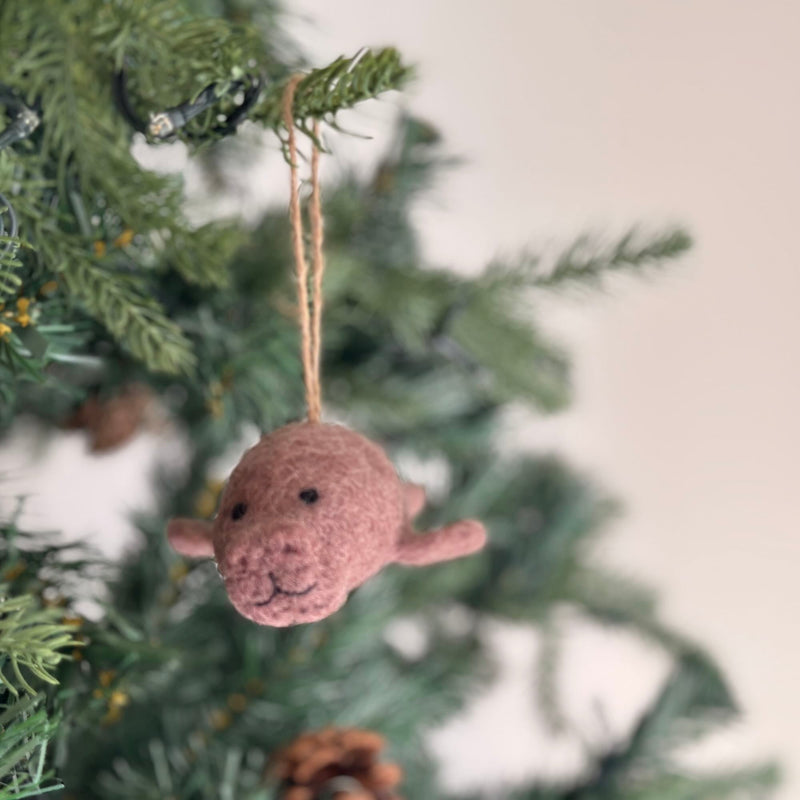A light pink handcrafted felt manatee ornament hangs gracefully from a festive Christmas tree branch. This unique felt manatee ornament features a rounded body, tiny flippers, and a simple stitched smile. A jute string allows it to blend into the green needles, subtle lights, and pine cones, creating a warm, inviting holiday atmosphere.
