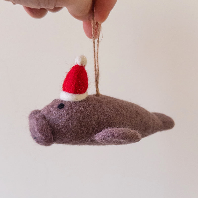 A light purple manatee felt ornament is held gently by a hand against a plain background. This charming Christmas tree ornament features a tiny black eye, a small round ear, and is adorned with a festive red and white Santa hat. Its soft felt texture is evident, and a jute string allows it to hang.