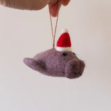 A muted purple wool felt manatee ornament is gently suspended by its jute string from a female hand against a soft, neutral backdrop. This handcrafted felt manatee Christmas ornament wears a festive red Santa hat trimmed with white felt and has small black eyes, exuding a whimsical and charming holiday spirit.
