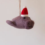 A handmade felt manatee ornament hangs, displayed against a light background. Crafted from soft, heathered-brown wool felt, this charming manatee features a rounded body, a flat tail, and small dark eyes. It wears a jaunty red felt Santa hat with a white brim and pompom, and a jute string is attached for hanging.
